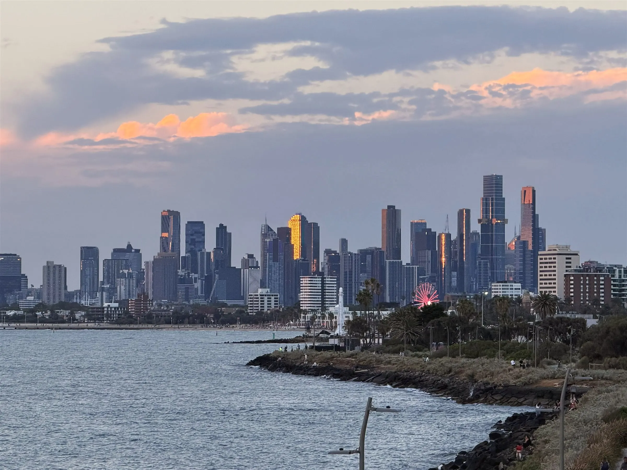 The Melbourne skyline across the bay at dusk, with pink clouds, towers catching the last light and a rocky foreshore in front