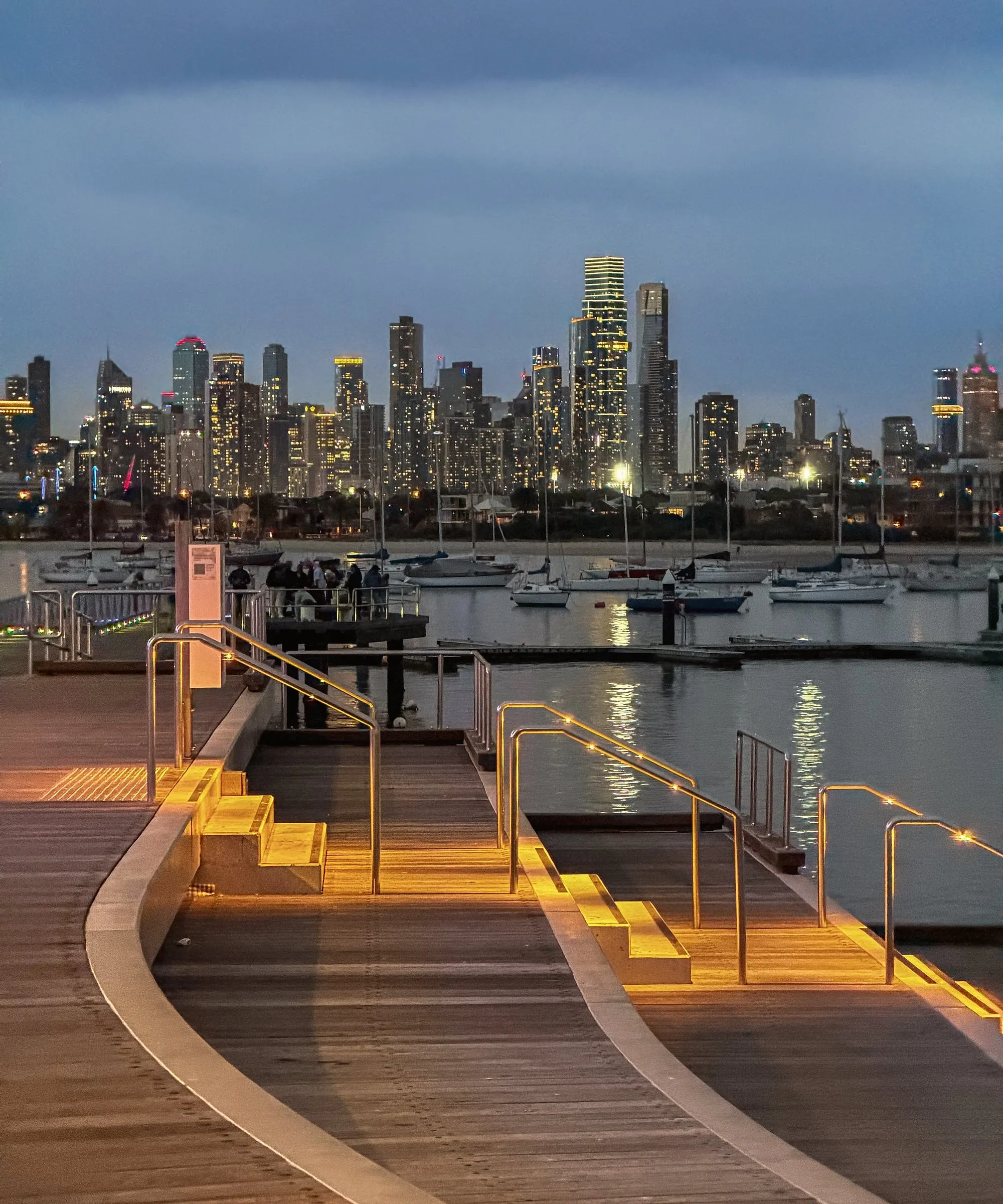 Timber steps lit with warm lights at dusk, with moored boats and the city skyline across the water