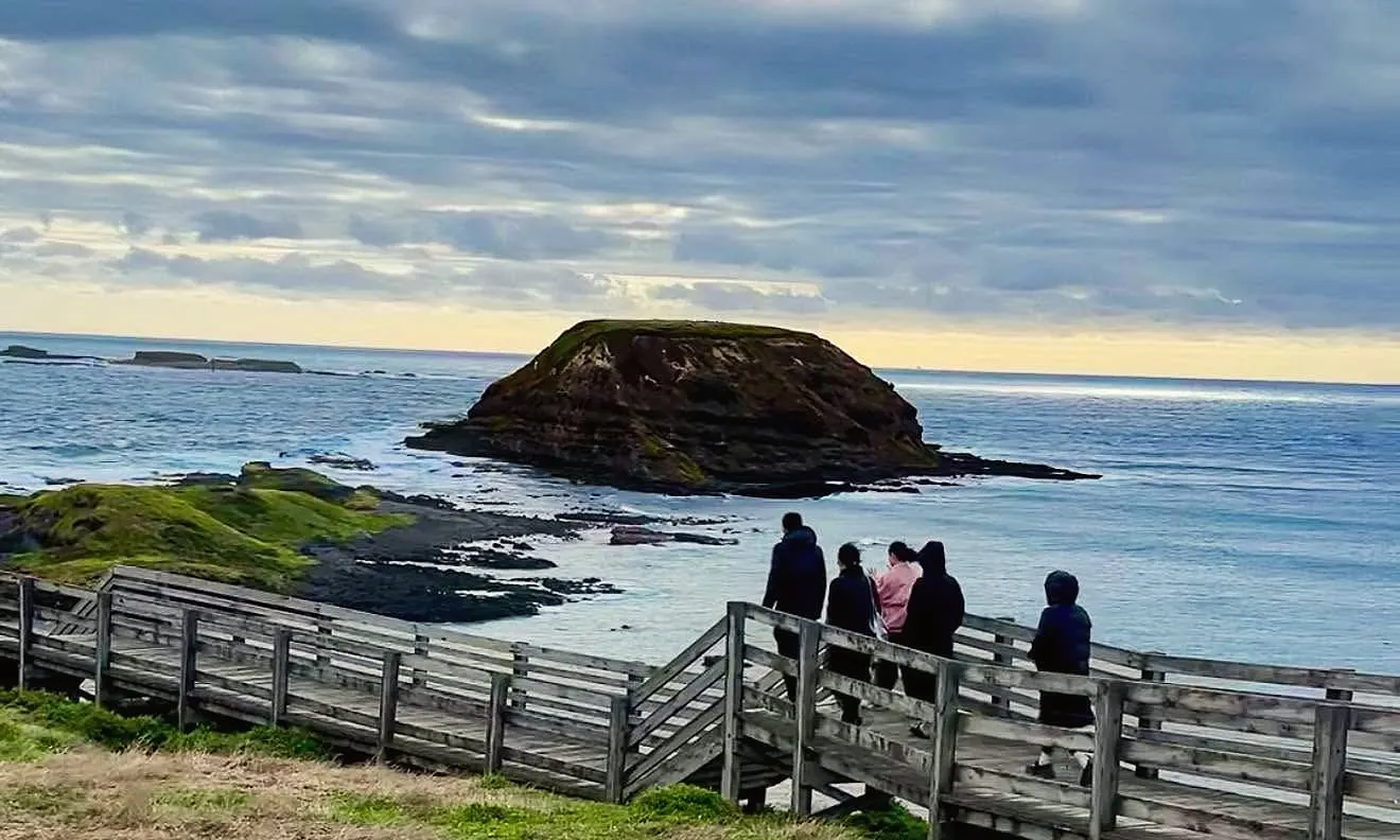 People on a timber boardwalk looking out at a rocky island in the sea under a soft, cloudy sky