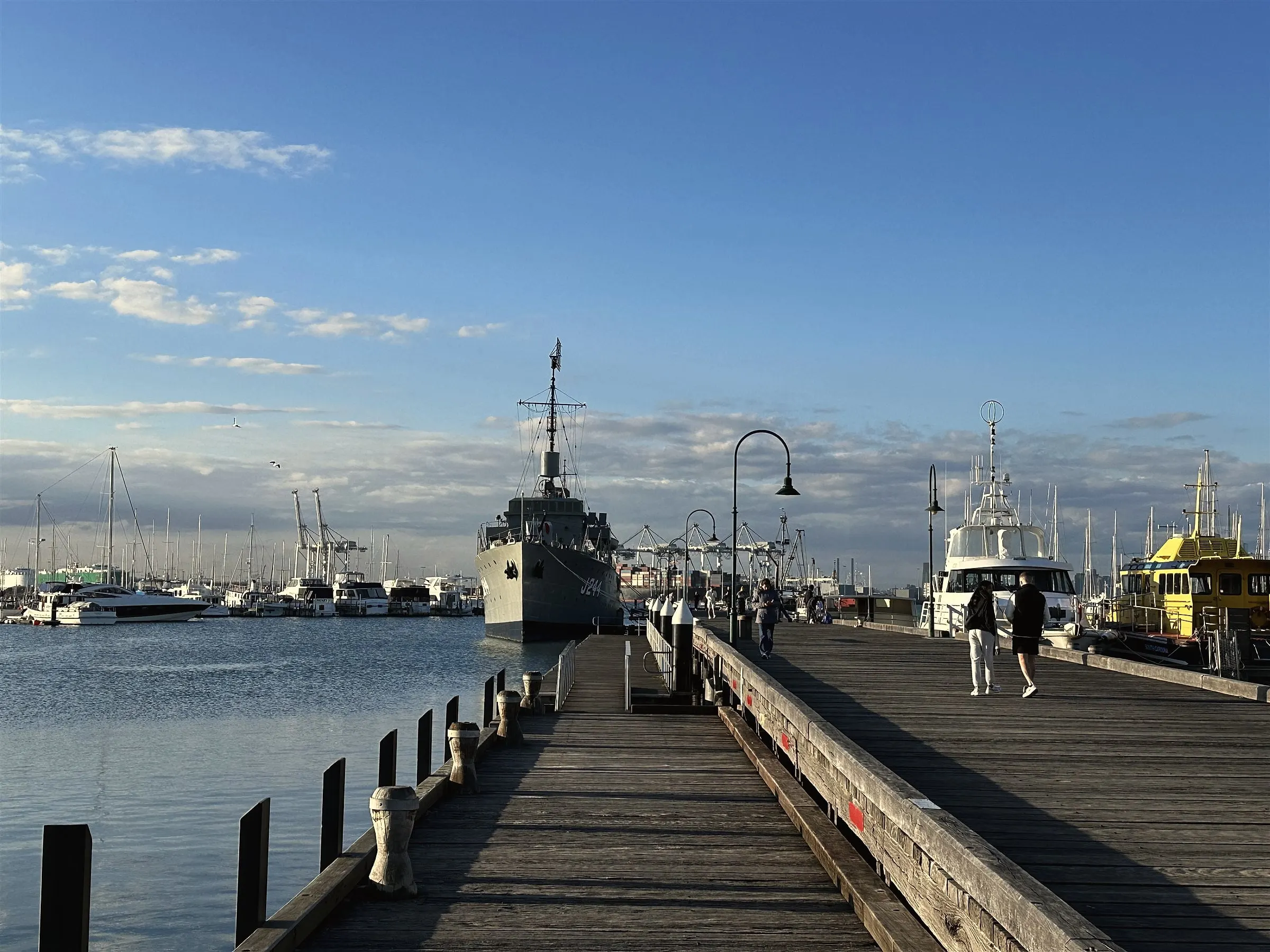 The grey former navy ship HMAS Castlemaine moored at the end of a timber pier, with boats on either side