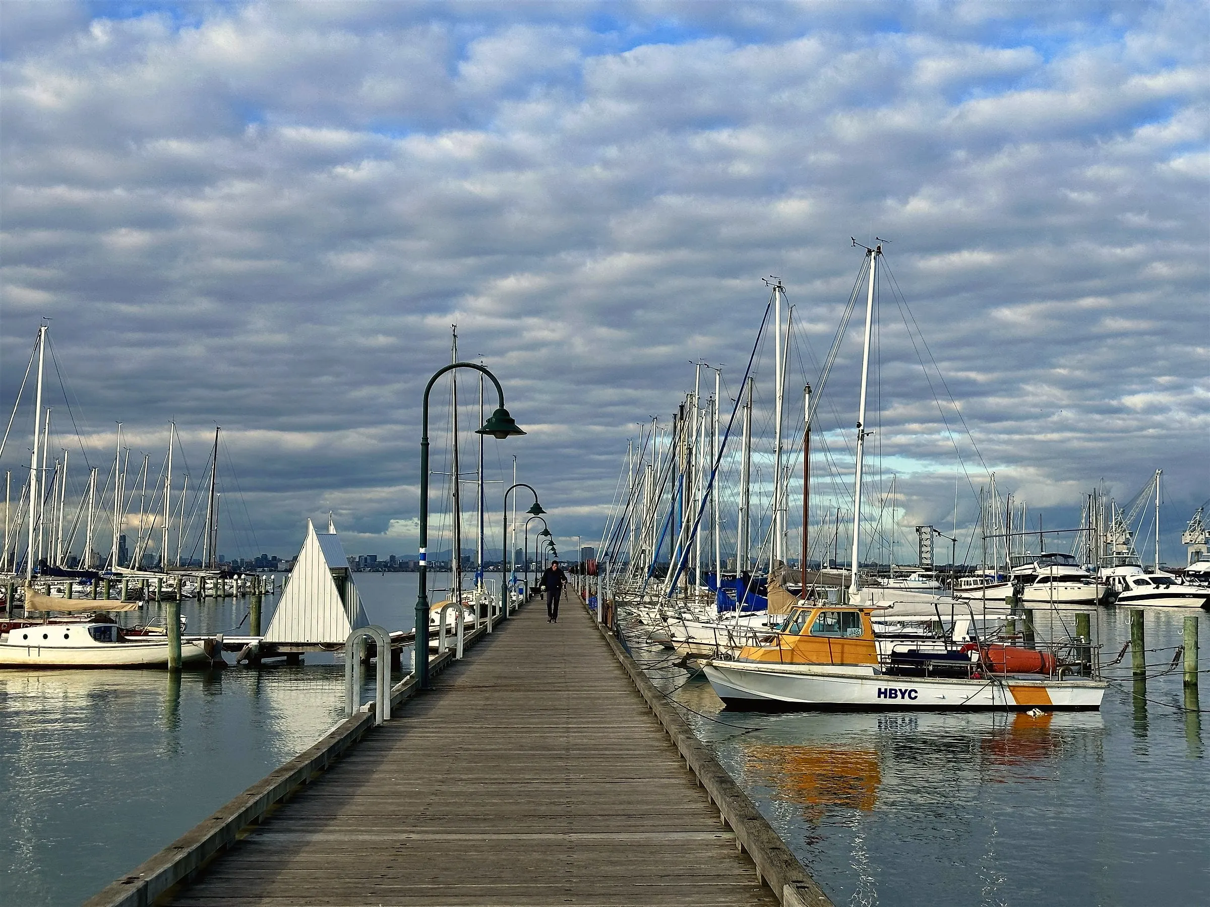 A long timber jetty running between moored yachts under a cloudy sky