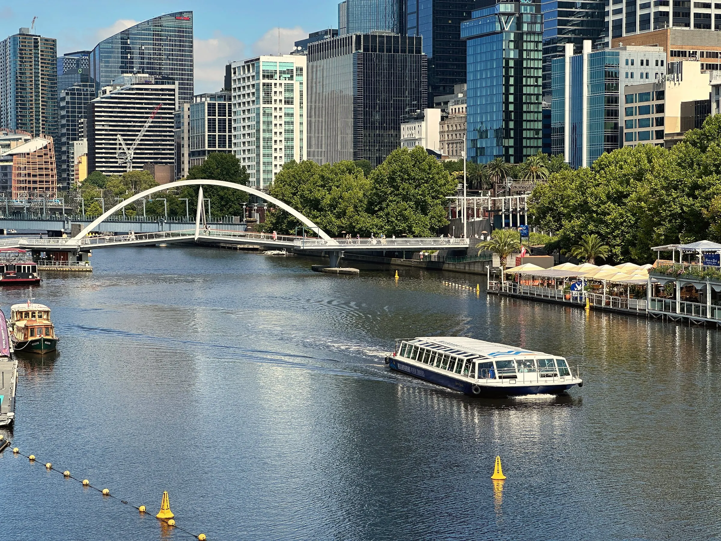 A river cruise boat on the Yarra, with an arched footbridge and city towers behind